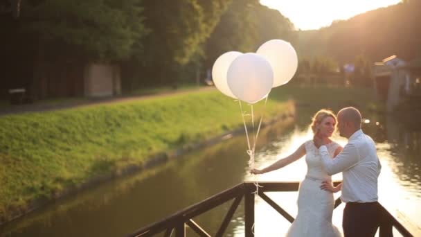 Tender moment van geluk. Jonge mooie bruidspaar met ballonnen omarmen op de rivier brug zonsondergang op de achtergrond. — Stockvideo