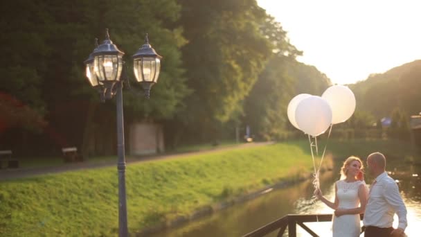 Moment tendre de bonheur et d'amour. Jeune beau couple de mariage avec des ballons embrassant sur le pont coucher de soleil rivière sur fond . — Video