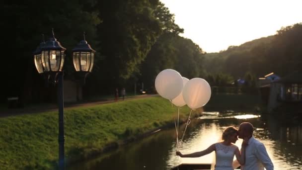 Momento terno de felicidade e amor. Silhoettes de jovem casal lindo casamento com balões abraçando na ponte do pôr do sol rio no fundo . — Vídeo de Stock