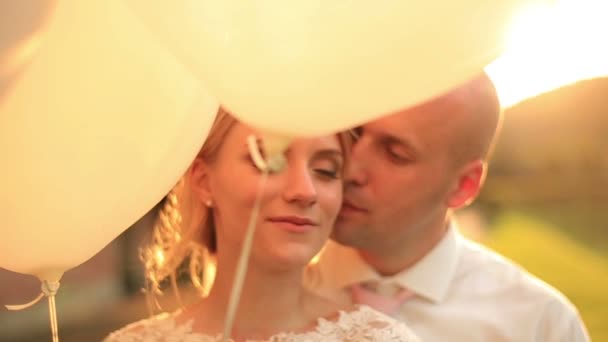 Glückliches junges Hochzeitspaar mit Luftballons bei Sonnenuntergang im Park. schöner Bräutigam küsst sanft seine schöne blonde Braut — Stockvideo