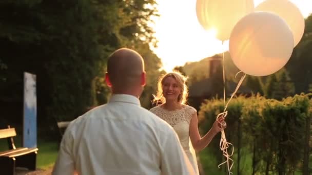 Lyckliga bruden och brudgummen promenader tillsammans och kyssas i gröna höst park håller vita ballonger i handen. Golden sunset bakgrund — Stockvideo