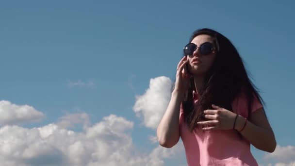 Young attractive brunette girl in sunglasses emotionally talking by phone against the blue cloudy sky — Stock Video