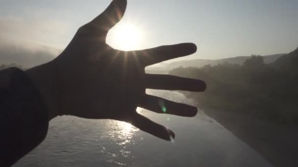 Primer plano de la mano de los hombres cubriendo el sol mientras amanece en el río de la montaña brumosa por la mañana — Vídeos de Stock