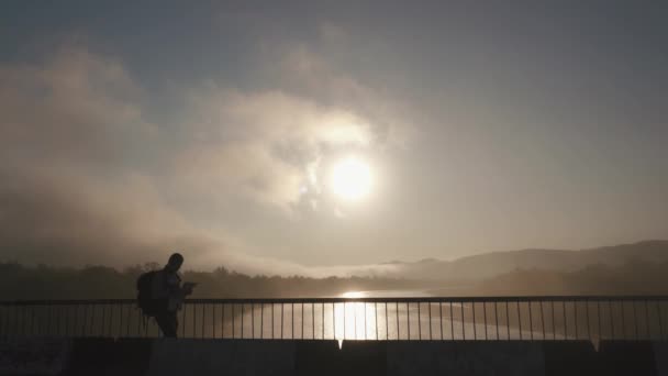Силуэт человека туриста в спортивной одежде с картой и рюкзаком прогуливается по мосту и пытается целиться в районы. Закат и облачный фон неба — стоковое видео