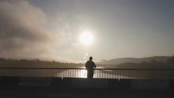 Silhouet van man toerist in sportkleding met kaart en rugzak doorloopt aan de brug wilt doelgebieden en horloges van de zonsondergang. Bewolkte hemelachtergrond — Stockvideo