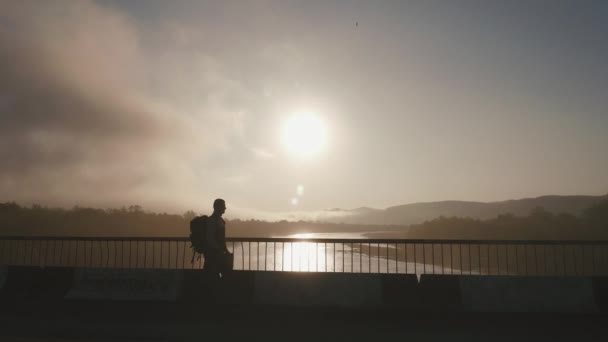 Silueta de hombre turista con mochila y mapa en sus manos en el puente. Maravilloso fondo atardecer — Vídeo de stock