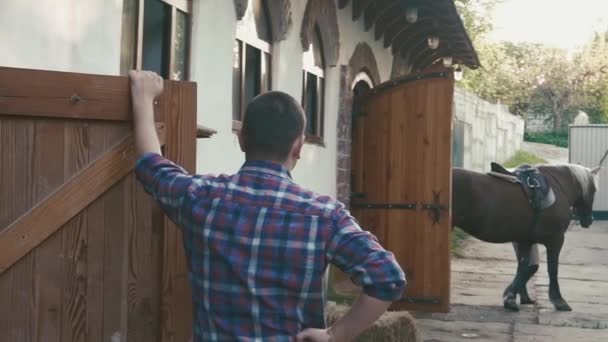 Young jockey girl is walking a horse out of a stable while trainer watches her from aside — Stock Video