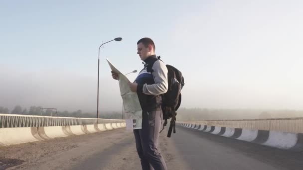 Gros plan de l'homme touriste en vêtements de sport avec carte et sac à dos marche à travers le pont et en essayant de cibler les zones — Video