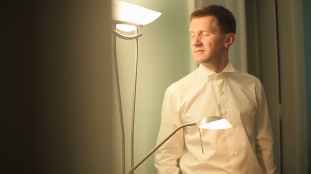 Feliz novio elegante preparándose para el día de la boda en la habitación del hotel. Hombre reflexivo con los ojos cerrados iluminado por la luz de la lámpara — Vídeo de stock