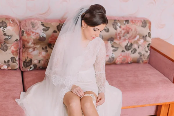 Beautiful bride sitting on sofa in dressing room is wearing bridal garter, preparing herself for wedding ceremony — Stock Photo, Image