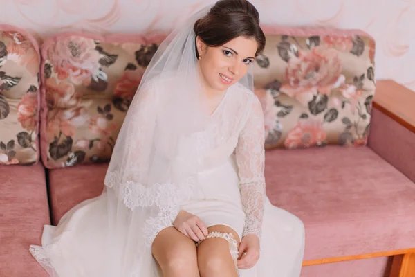 Encantadora novia sentada en el sofá en el vestidor está usando liga nupcial, preparándose para la ceremonia de boda —  Fotos de Stock