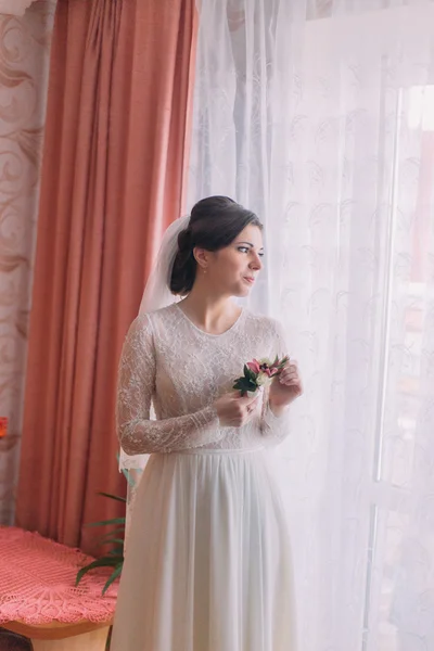 Retrato de media longitud de novia encantadora en vestido de novia cerca de la ventana que sostiene boutonniere — Foto de Stock