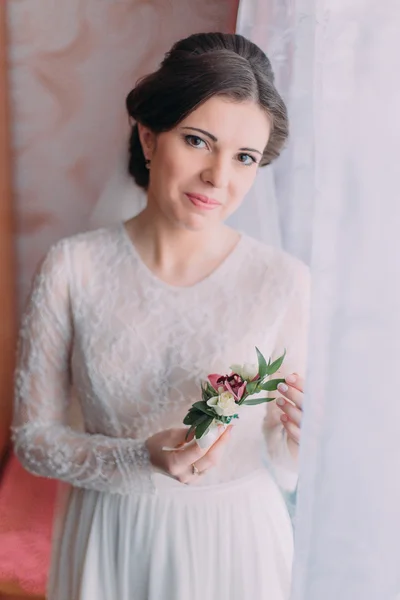 Retrato de meio comprimento de noiva bonita em vestido de noiva perto de janela brilhante interior segurando boutonniere — Fotografia de Stock