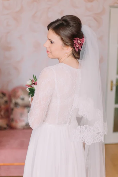 Meia-comprimento retrato de noiva bonita em vestido de noiva olhando sobre seu ombro dentro de casa segurando boutonniere — Fotografia de Stock