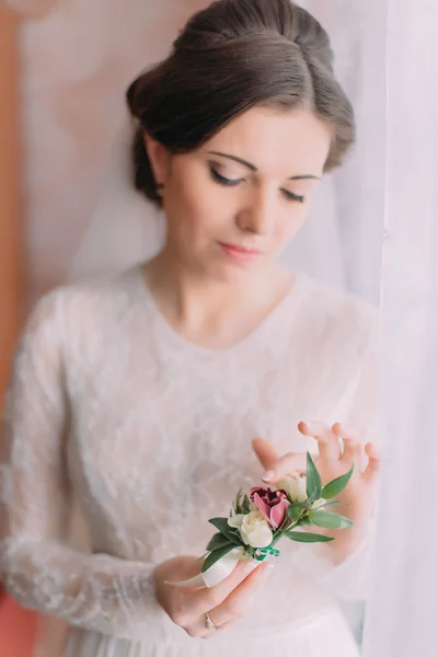 Foto close-up pengantin wanita cantik yang tidak bersalah dalam gaun pengantin dekat jendela memegang boutonniere lucu — Stok Foto