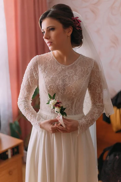 Ritratto di mezza lunghezza di bella sposa in abito da sposa posa al chiuso tenendo boutonniere poco floreale — Foto Stock