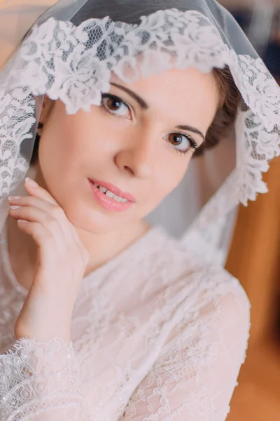 Beautiful sensual bride in wedding gown with lifted veil touching her face while posing indoors — Stock Photo, Image