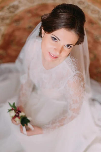 Close-up retrato de bela noiva sorridente em vestido de noiva sentado no tapete e segurando bonito boutonniere floral — Fotografia de Stock
