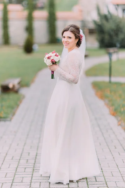 Чувственная молодая красивая невеста, держащая свадебный букет, пока позирует на дорожке парка в солнечный день — стоковое фото