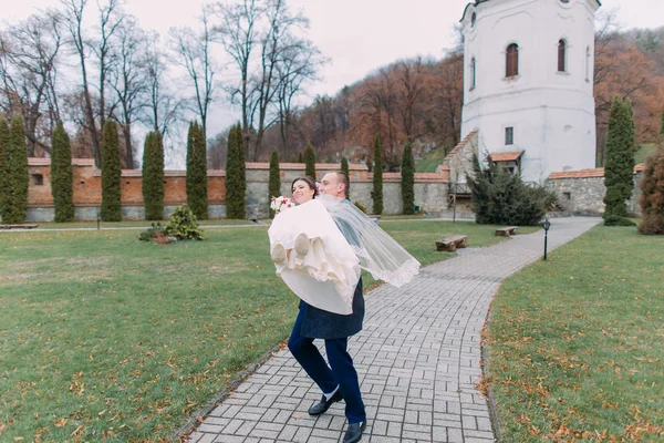Страстная молодожёны в день свадьбы прогуливаются по красивому зеленому парку. Любящий жених держит свою новую жену на руках — стоковое фото