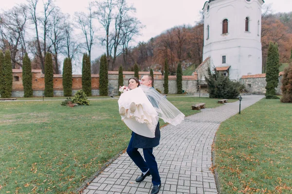 Passionate newlywed couple at wedding day walking outdoors beautiful green park. Loving groom hold his new wife on hands — Stock Photo, Image