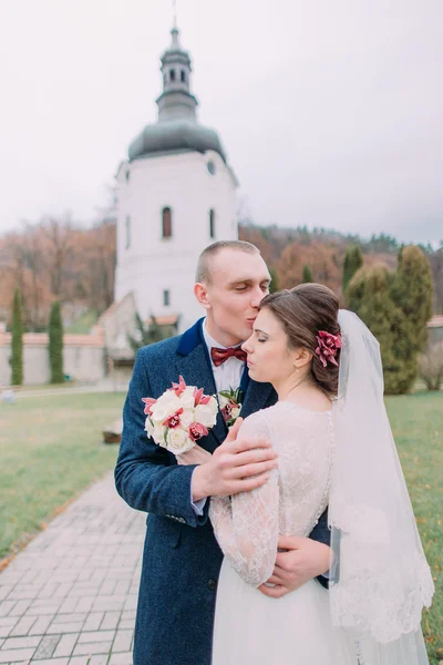 Красивий наречений ніжно обіймає і цілує свою нову дружину в зеленому парку старого монастиря — стокове фото