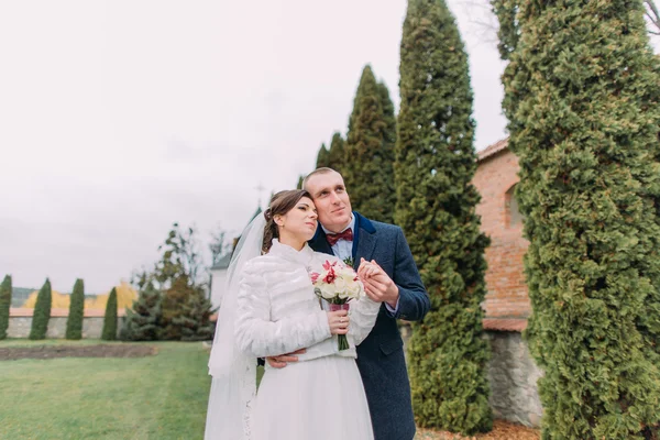 Schöner Bräutigam hält sanft Hand seiner eleganten neuen Frau, während beide von ihrem Eheleben in der Nähe von Zypressenreihen im grünen Park träumen — Stockfoto