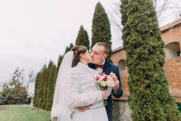 Guapo novio tiernamente besando a su elegante nueva esposa cerca de los cipreses y la pared fortificada en el parque romántico —  Fotos de Stock