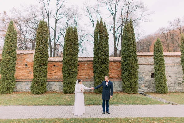 Красивый жених позирует со своей женой возле кипарисов и укрепленной стены в романтическом парке — стоковое фото