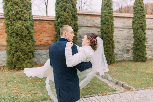 Красивий наречений тримає свою нову дружину на руках біля кипарисів і укріпленої стіни в романтичному парку — стокове фото