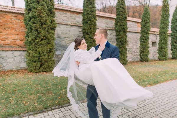 Handsome groom holding his elegant bride on hands near cypress trees and fortified wall in romantic park — Stock Photo, Image