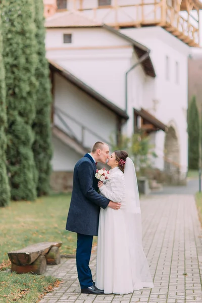 Красивий наречений ніжно цілує свою ніжну наречену на прогулянці в зеленому парку з древніми будівлями і кипарисами — стокове фото