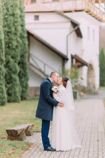 Bello sposo che abbraccia teneramente la sua tenera sposa a passeggiare nel parco estivo con edifici antichi e cipressi verdi — Foto Stock