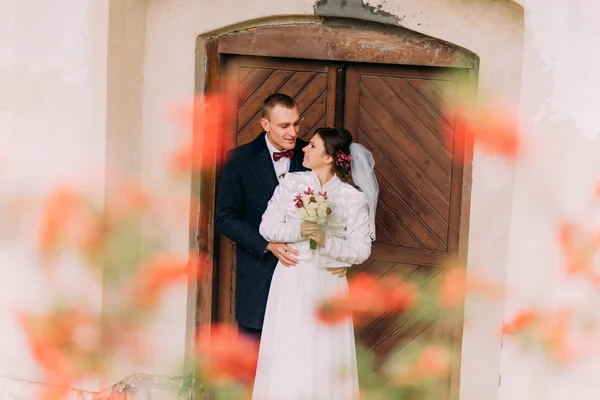 Stilig nygifta par kramar nära ingången av antika förstörda slott med söta små röda blommor på förgrunden — Stockfoto