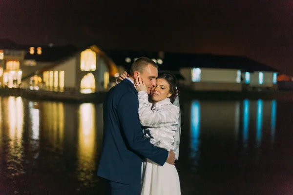 Retrato romántico de boda. Novia y novio posando cerca del lago nocturno iluminado con luz brillante de las ventanas de la sala de banquetes — Foto de Stock
