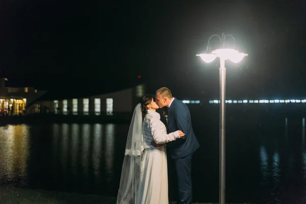 Matrimonio romantico. Allegri sposi che si baciano vicino al lago notturno illuminato con luce brillante dalle finestre della sala banchetti — Foto Stock