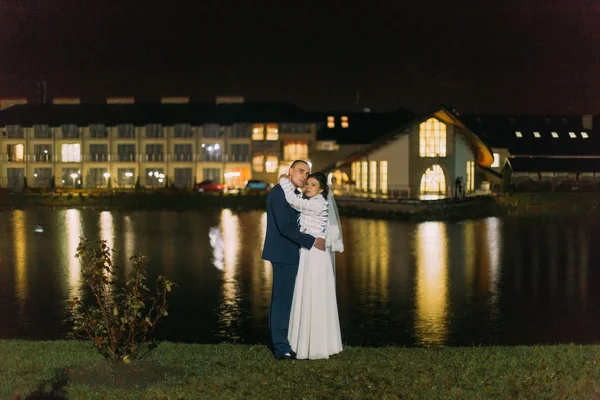 Романтический свадебный портрет. Возбужденные невеста и жених позируют возле ночного озера, освещенного ярким светом из окон банкетного зала — стоковое фото