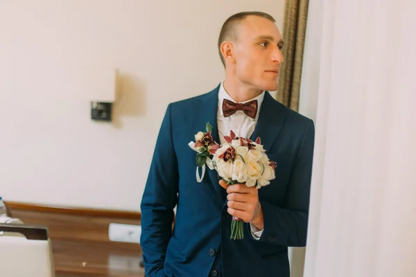 Elegante hombre vestido sosteniendo elegante ramo floral de pie cerca de la pared de la habitación de hotel —  Fotos de Stock