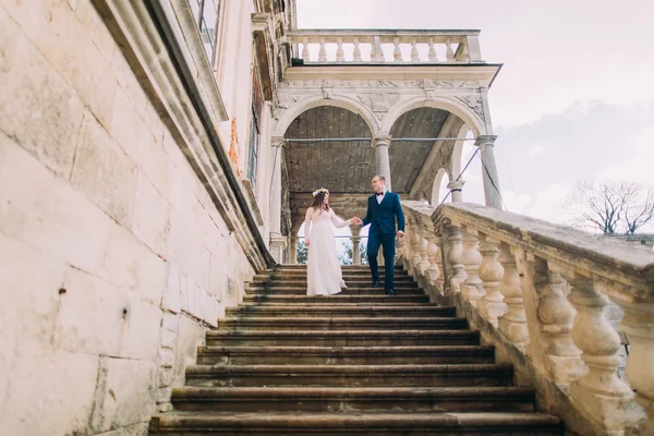 Suave novio cogido de la mano de su novia bonita al descender por escaleras de piedra palacio antiguo. Disparo de ángulo bajo — Foto de Stock