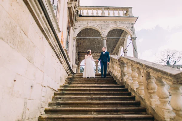 Der sanfte Bräutigam hält Händchen, wenn er mit seiner hübschen Braut die Steintreppe des antiken Palastes hinuntergeht. Flachschuss — Stockfoto