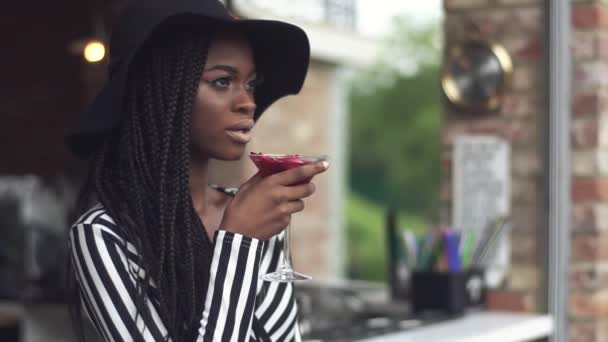 Шикарная африканская деловая леди в черной шляпе пьет коктейль в баре и наслаждается своим напитком — стоковое видео