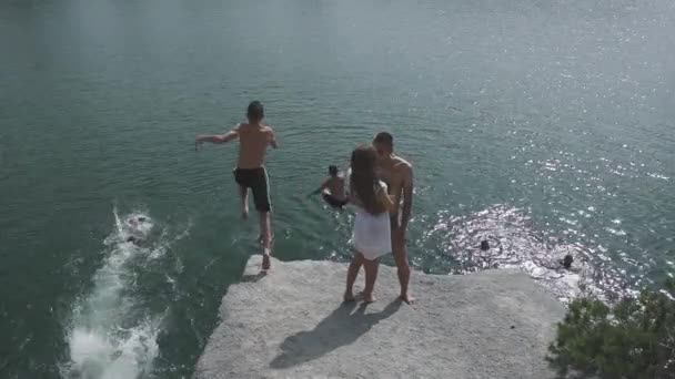 Ein verliebtes Teenie-Paar küsst sich auf einem großen Stein in der Nähe eines großen Sees, während andere junge Leute springen und baden. Zeitlupe — Stockvideo