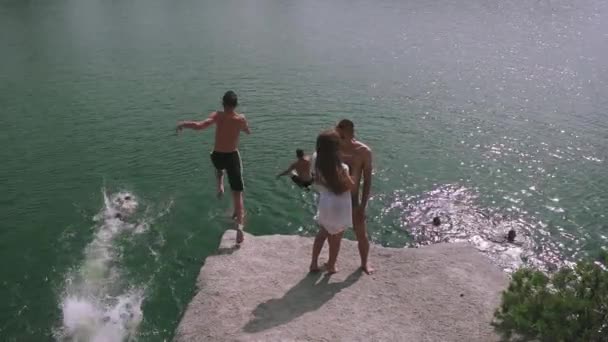 愛するティーンエイジャーのカップルは、他の若者がジャンプして入浴して水面近くの大きな石にキスをしています。スローモーション — ストック動画
