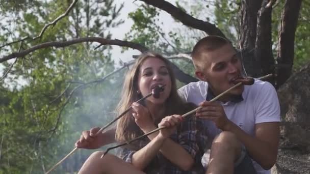 Heureux couple d'adolescents mangent des saucisses frites sur un feu de joie fumé dans la forêt lors d'une randonnée — Video