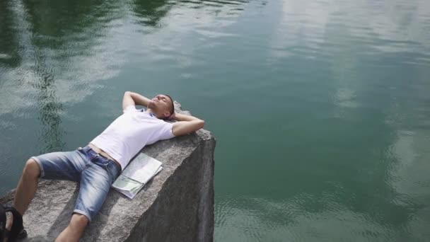 Jovem homem ativo relaxa descansando na rocha perto da superfície calma lago montanha com mapa que coloca ao lado dele — Vídeo de Stock