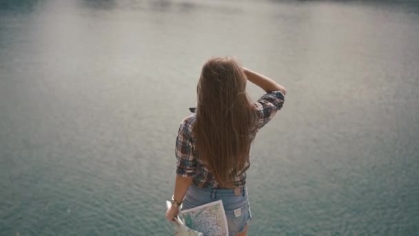 Beautiful young active backpacker girl checking with map near water surface of mountain lake at sunny day — Stock Video