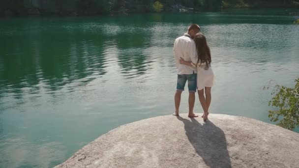 Hermosa pareja amada besándose al aire libre en la orilla del lago de pie sobre una roca grande — Vídeo de stock