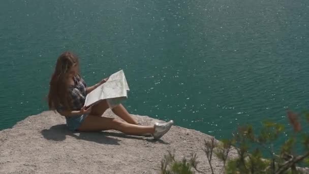 Молодая активная девушка отдыхает на горной скале на большой водной поверхности горного озера и ищет карту — стоковое видео