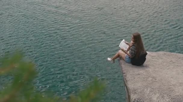 Young active backpacker girl resting on mountain rock upon large water surface of mountain lake and looking to map — Stock Video