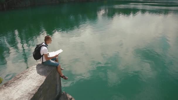 Jeune homme actif se détend sur la pente rocheuse sur la surface calme du lac de montagne lecture carte papier dans ses mains — Video